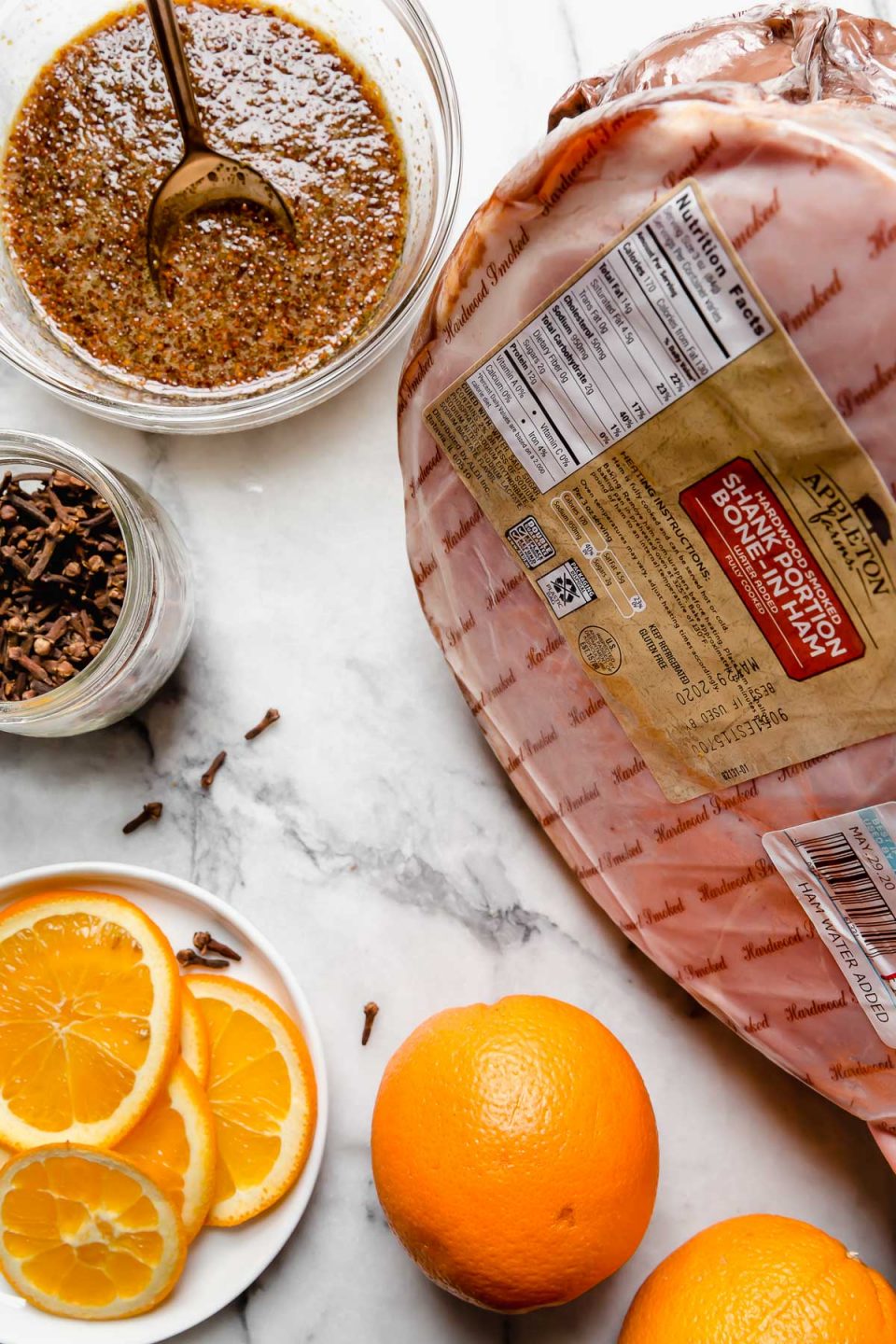 Ingredients for this maple glazed ham recipe on a white marble surface: smoked ham, oranges, cloves & maple glaze.
