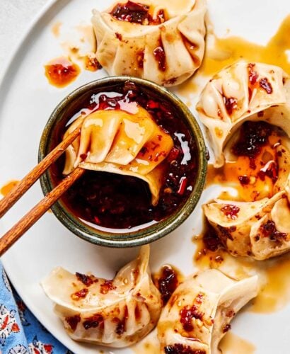 An overhead shot of a pair of chopsticks dunking a dumpling into a dish of dumpling sauce on a white plate with several more dumplings.