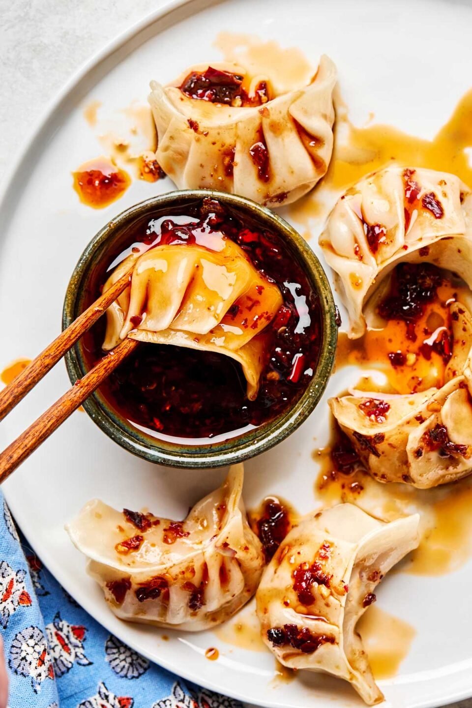 An overhead shot of a pair of chopsticks dunking a dumpling into a dish of dumpling sauce on a white plate with several more dumplings.