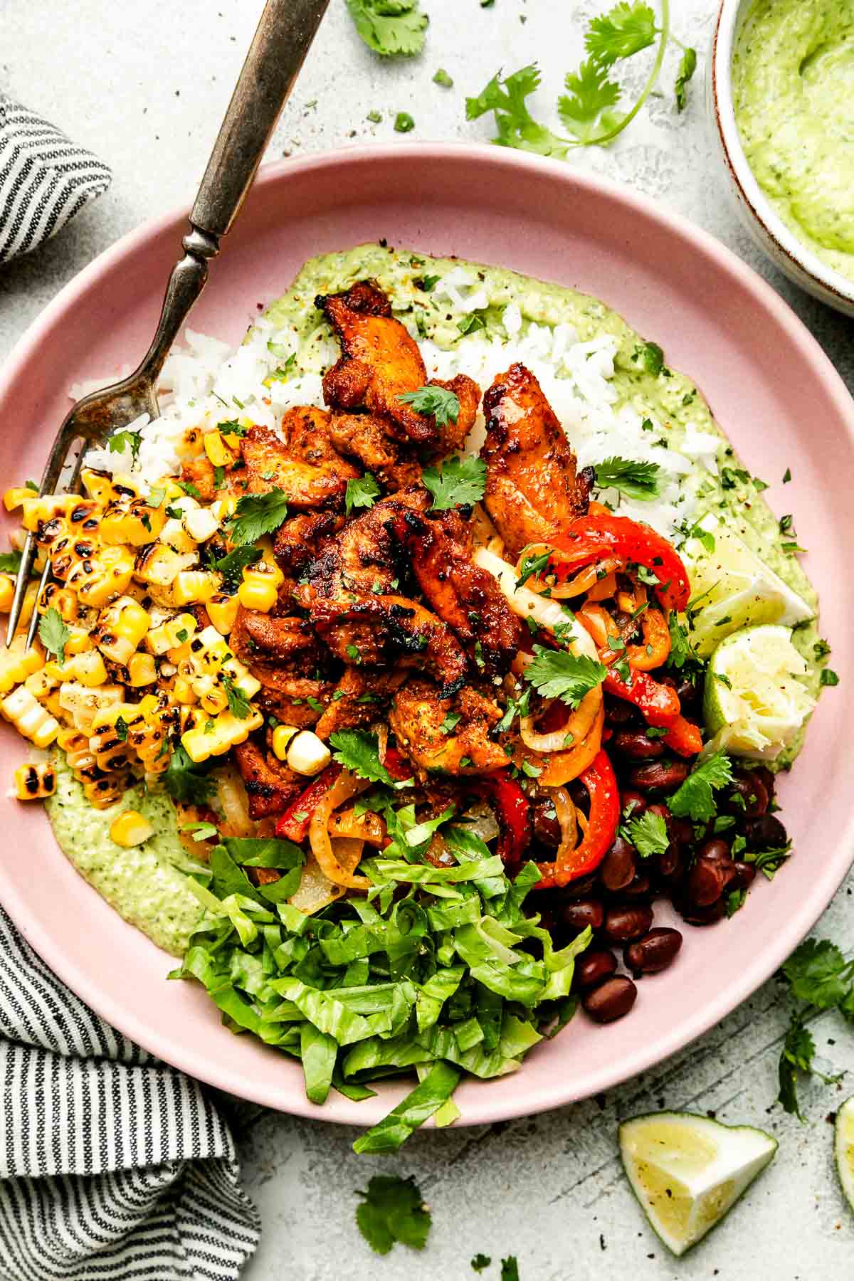 An overhead shot of an assembled fajita bowl on a light pink plate alongside a striped cloth on a light grey surface: avocado crema, fajitas, lettuce, corn, pico de gallo, rice and black beans, topped with cilantro.