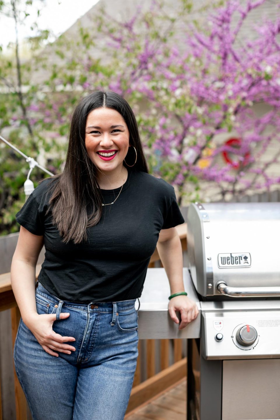 Jess of Plays Well With Butter stands on a deck in front of a stainless steel Weber Genesis II propane grill. In the background, a blossoming tree & a house with beige siding.