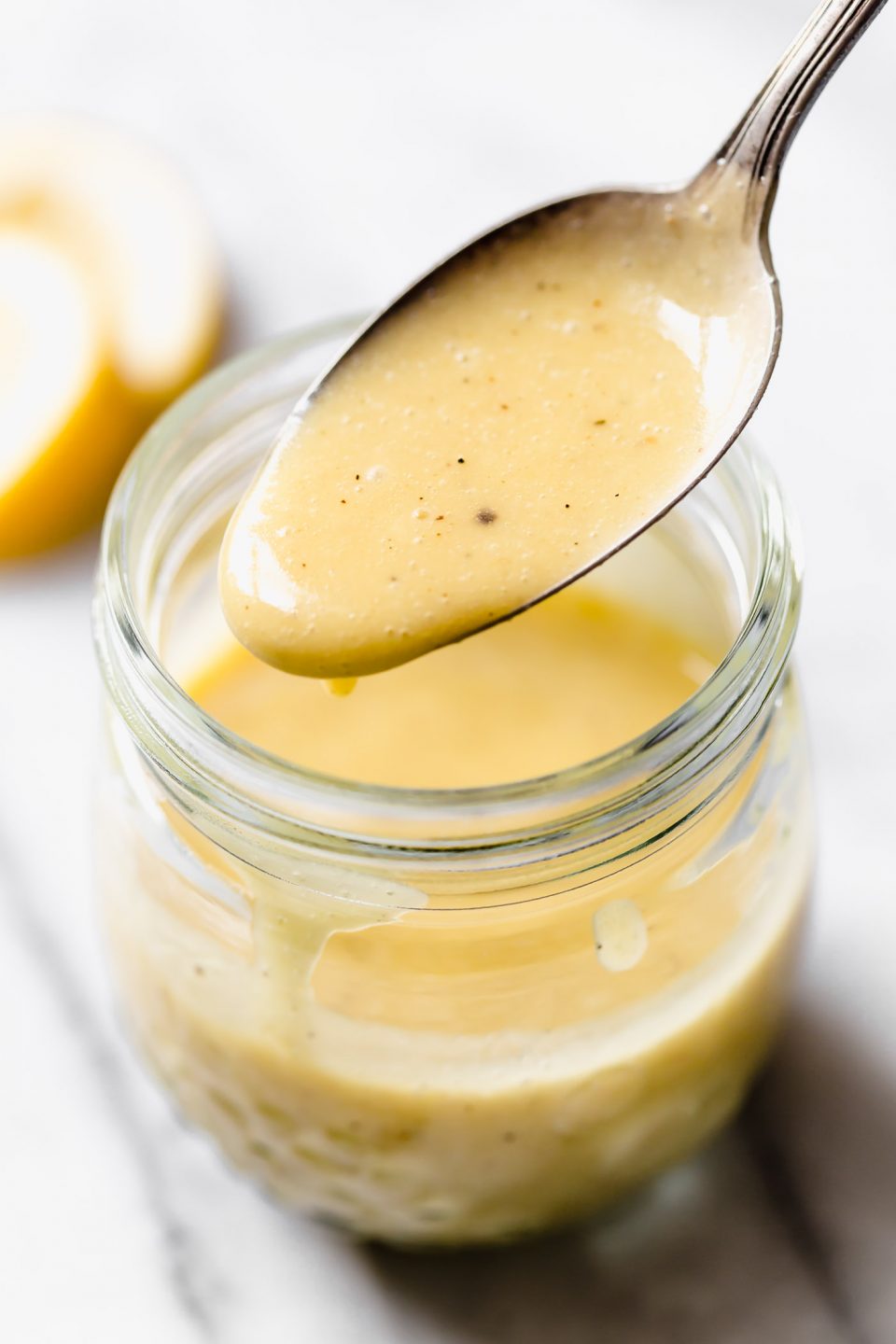 All purpose vinaigrette in jar with spoon drizzling vinaigrette back into jar. Set on white marble backdrop. Lemons in the background.