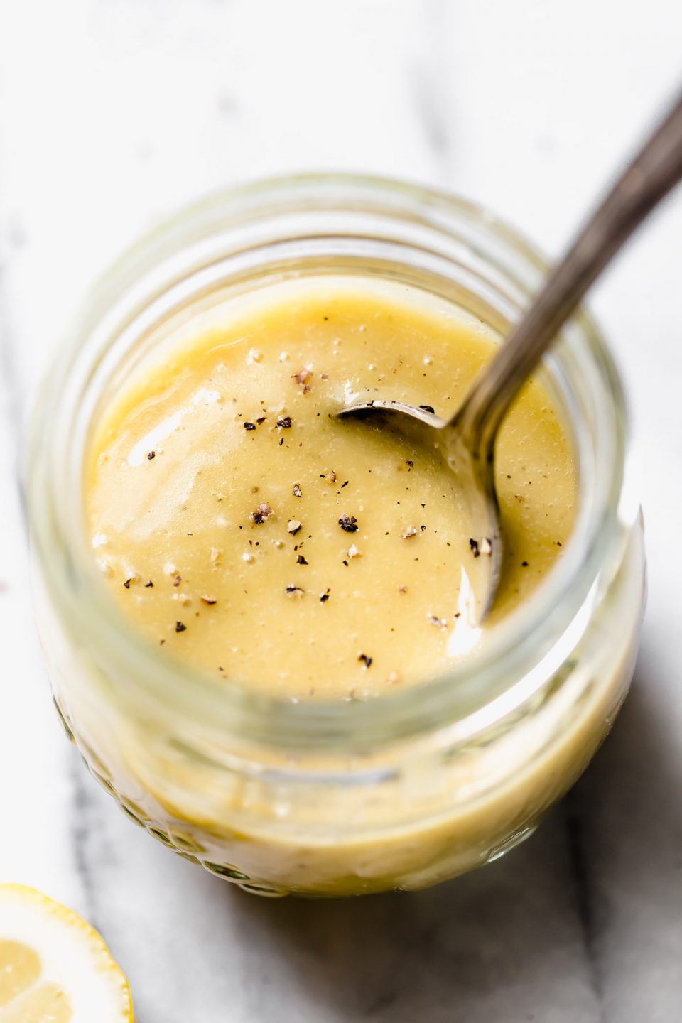 All purpose vinaigrette in jar set on white marble backdrop. Spoon coming out of jar. Cracked black pepper on surface of salad dressing. Lemons in the background.