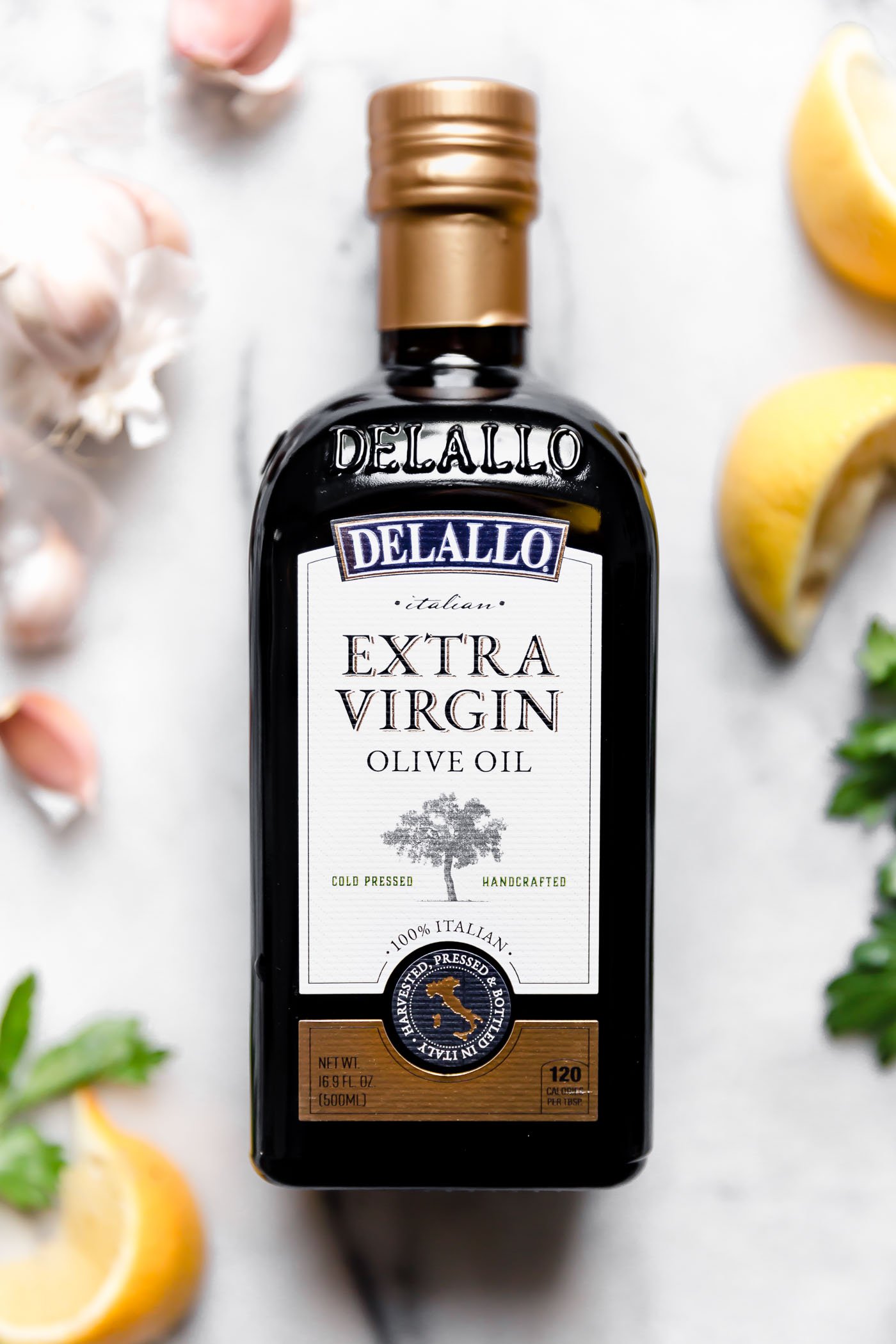 DeLallo olive oil bottle shown on a white marble surface surrounded by lemon wedges, whole garlic cloves & parsley.