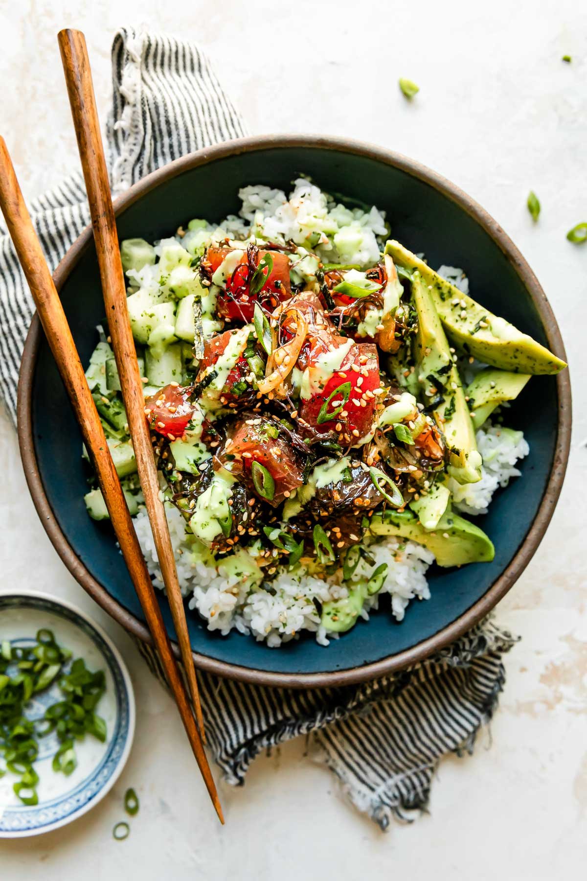 An overhead shot of an ahi tuna poke bowl assembled inside of a large blue ceramic bowl. The bowl sits atop a creamy white textured surface and a pair of wooden chop sticks resting atop the bowl. A blue and white linen napkin is tucked underneath the bowl and a small blue and white plate filled with sliced green onions rests alonside the bowl at center.