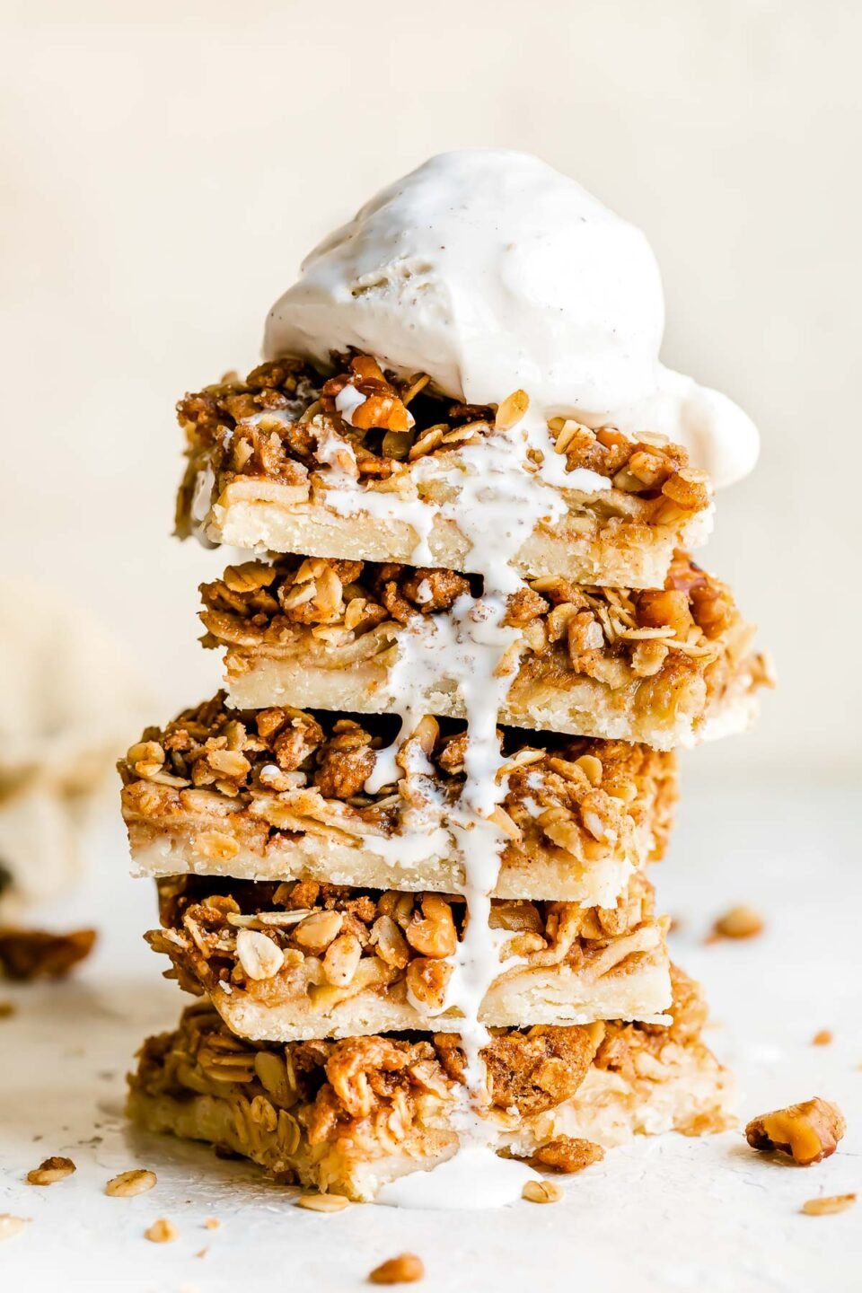 A side angle shot of five apple pie bars stacked on top of each other served with a scoop of vanilla ice cream melting over top. The bars and ice cream are stacked atop a creamy white textured surface.