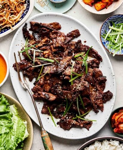 An overhead shot of a white platter of beef bulgogi alongside dishes of rice, green onions, gochujang aioli, cucumber kimchi, mung beans, kimchi and leafy lettuce.