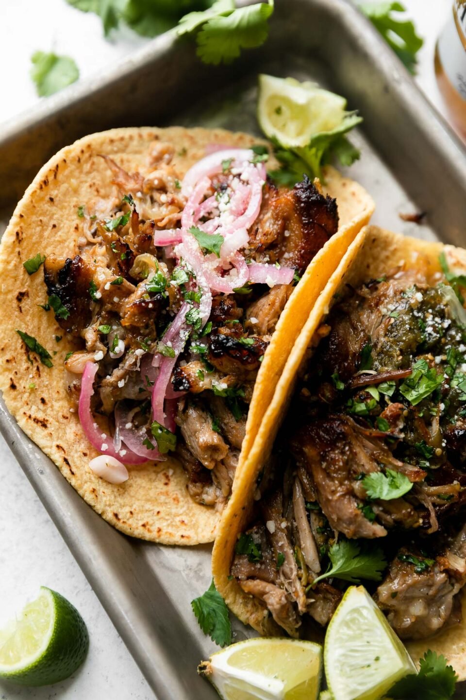 A side angle shot of beer braised carnitas tacos topped with pickled red onions, fresh chopped cilantro, queso fresco, with lime wedges on the side. The tacos sit atop a parchment paper lined aluminum baking sheet that sits atop a creamy white surface surrounded by lime wedges, a bouquet of fresh cilantro, and a bottle of Modelo beer.