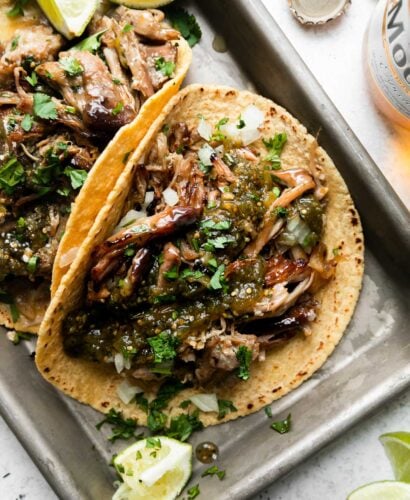 An overhead shot of beer braised carnitas tacos topped with salsa verde, fresh chopped cilantro, diced yellow onion, with lime wedges on the side. The tacos sit atop an aluminum baking sheet that sits atop a creamy white surface surrounded by lime wedges, a bottle of Modelo beer, and a loose beer bottle cap.