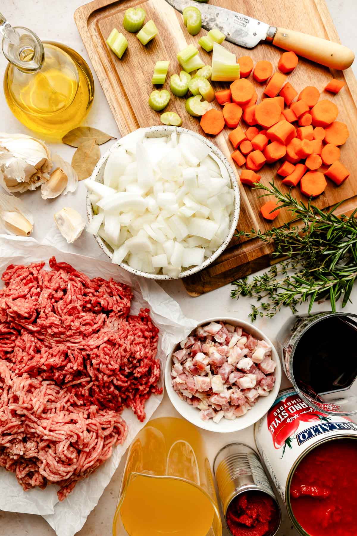 Bolognese sauce ingredients arranged on a creamy white textured surface: carrots, celery, yellow onion, pancetta, garlic, olive oil, ground beef, ground pork, tomato paste, red wine, fresh rosemary, fresh thyme, bay leaves, crushed tomatoes, low-sodium chicken broth or water, heavy cream, grated parmesan.