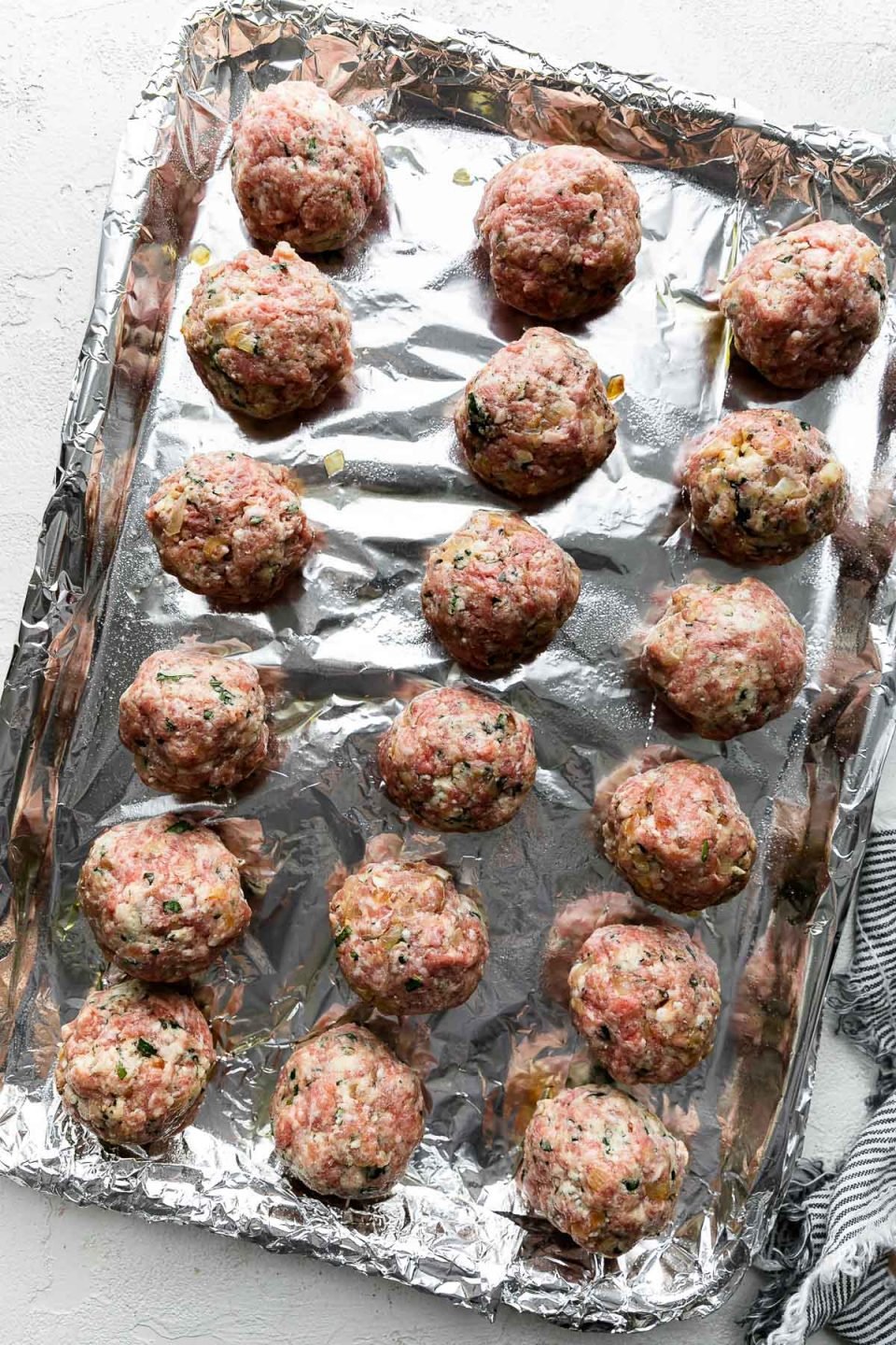 18 uncooked golf ball sized meatballs are arranged atop an aluminum foil lined baking sheet. The baking sheet rests atop a creamy white cement surface. A blue and white striped linen napkin rests alongside the baking sheet.