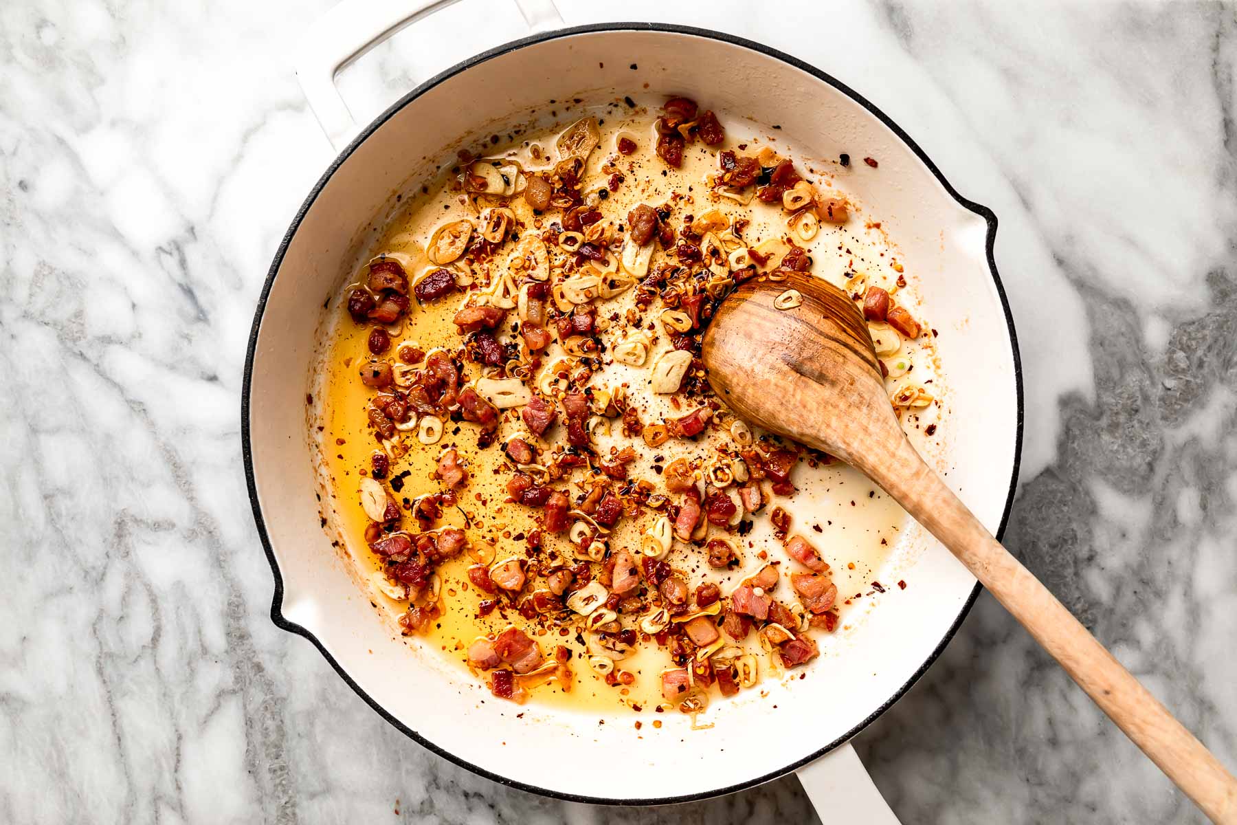 An overhead shot of pancetta and aromatics simmering in olive oil in a white skillet atop a white marbled surface.