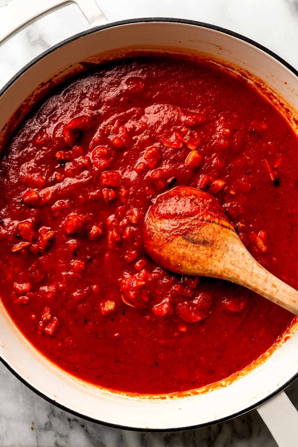 An overhead shot of tomato sauce in a white skillet with a wooden spoon, sitting atop a white marbled surface.
