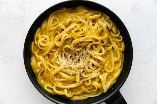 A small skillet filled with cooked linguine noodles tossed in creamy butternut squash pasta sauce. The noodles have been garnished with freshly grated parmesan & ground black pepper. The skillet sits atop a white textured surface.