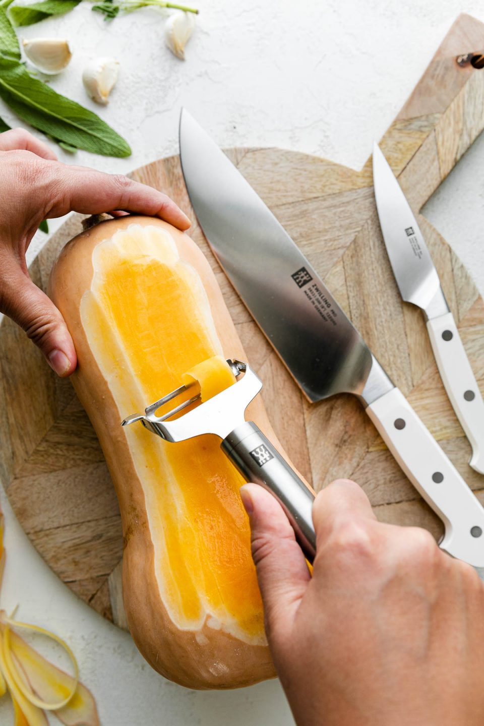 A whole butternut squash rests on top of a round wooden cutting board alongside a pair of Zwilling Pro Le Blanc Knives. A woman's hand holds the top portion of the butternut squash, while holding a Zwilling Pro Y-Peeler in the other hand to peel the squash. The cutting board sits atop a white textured surface while butternut squash peelings, fresh herbs, and cloves of garlic surround the cutting board.