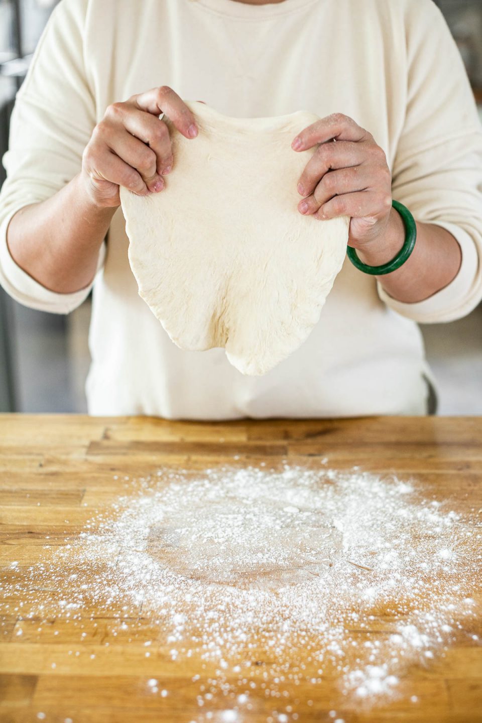 How to form pizza crust: Jess, shown in a white sweatshirt, stands behind a butcher block counter dusted in flour. Jess is holding the edges of pizza crust, stretching it out with her hands.