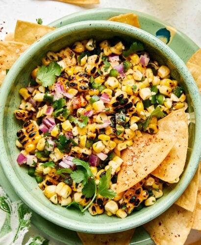 An overhead shot of sweet corn salsa with two tortilla chips in a light green bowl atop a matching plate full of chips. Extra chips, a margarita and a green and white striped cloth napkin sit alongside the plate.