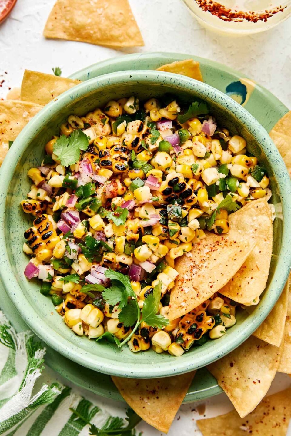 An overhead shot of sweet corn salsa with two tortilla chips in a light green bowl atop a matching plate full of chips. Extra chips, a margarita and a green and white striped cloth napkin sit alongside the plate.