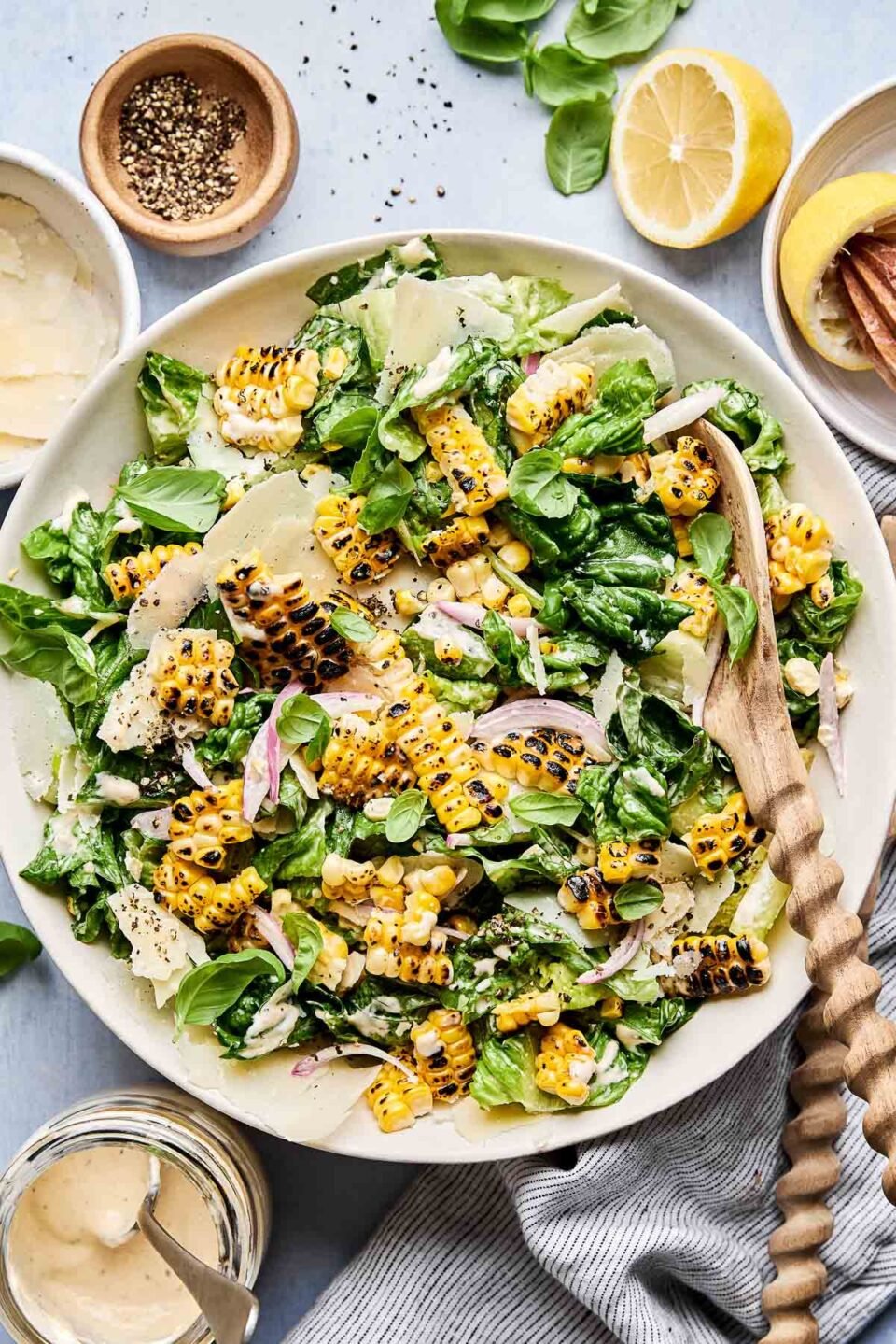 An overhead shot of charred sweet corn Caesar salad in a large shallow white bowl with a wooden serving spoon alongside a striped cloth on a light blue surface. The bowl is also surrounded by halved and juiced lemons, a jar of dressing, and bowls of black pepper and parmesan cheese.