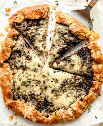 An overhead shot of a baked French onion galette with three sliced wedges on a piece of parchment paper atop an off-white surface. A spatula is under one of the slices to lift it.