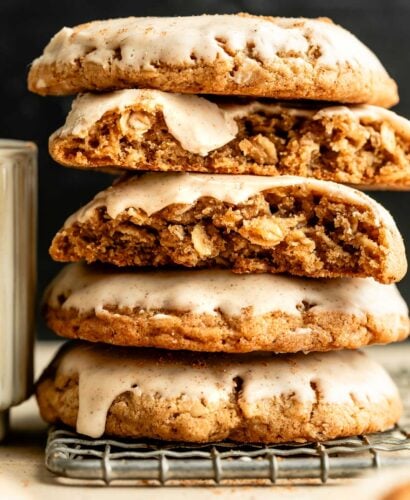 A side shot of five glazed oatmeal cookies stacked on a drying rack on a tan surface. Two of the cookies have a piece broken off, showing the interior texture.