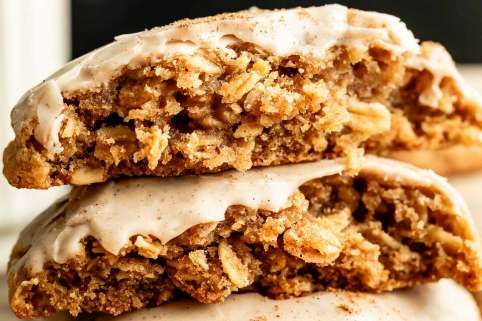 A side shot of three glazed oatmeal cookies stacked on a drying rack on a tan surface. Two of the cookies have a piece broken off, showing the interior texture. Glaze drips down from the bottom cookie onto a baking sheet.