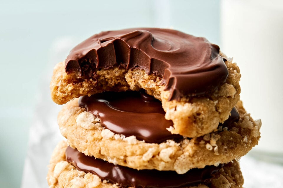 A close-up of three stacked oatmeal cookies topped with a layer of chocolate, with the top cookie partially bitten. A glass of milk is blurred in the background.