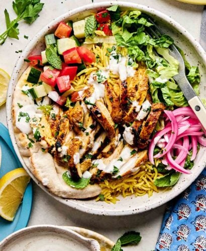 An overhead shot of a chicken shawarma bowl on an off-white surface alongside a patterned blue cloth, lemon wedges, shawarma sauce, and fresh herbs.