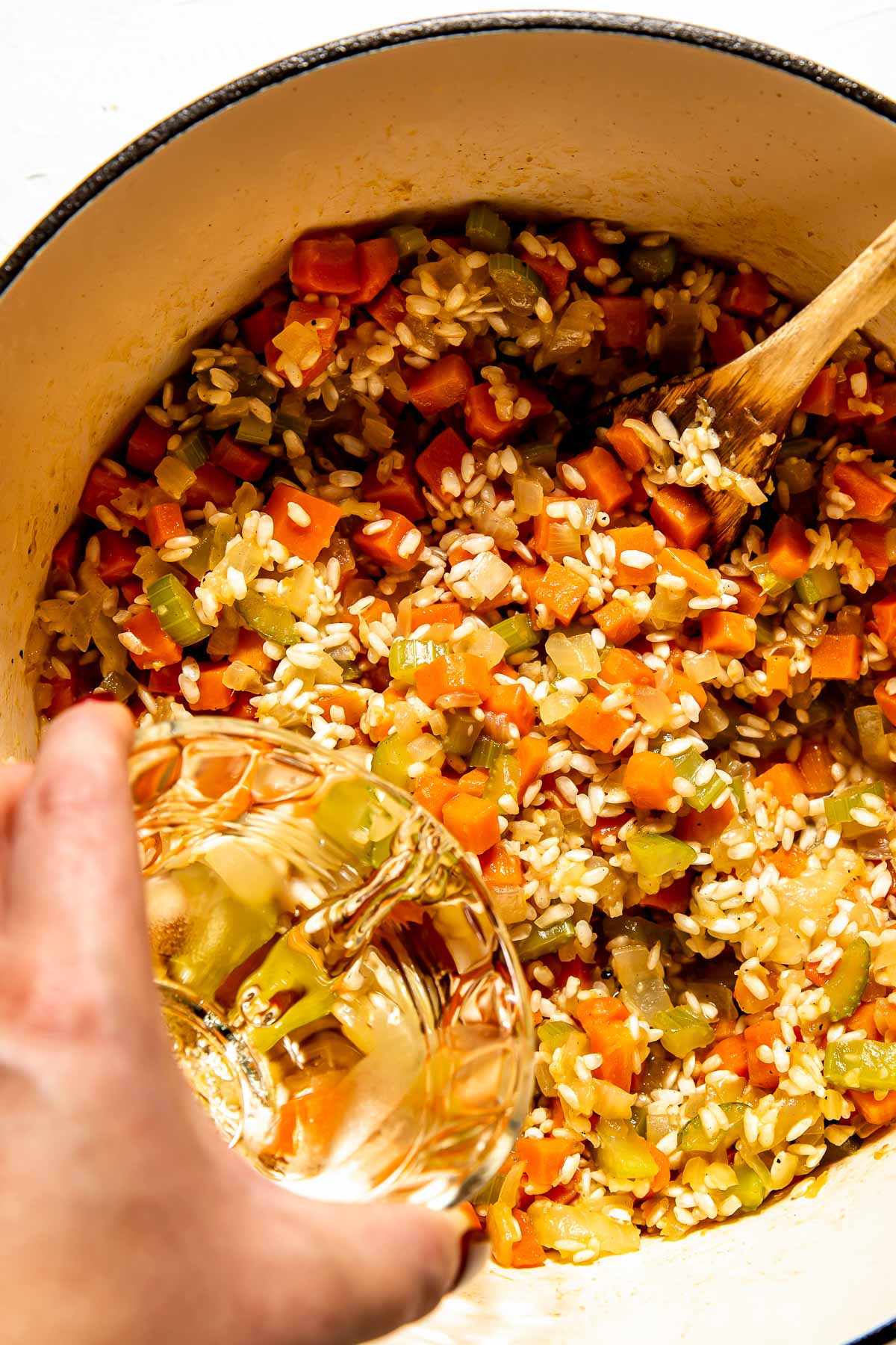 An overhead shot of a woman's hand pouring white wine over rice and softened vegetables in a large white pot atop a white surface.