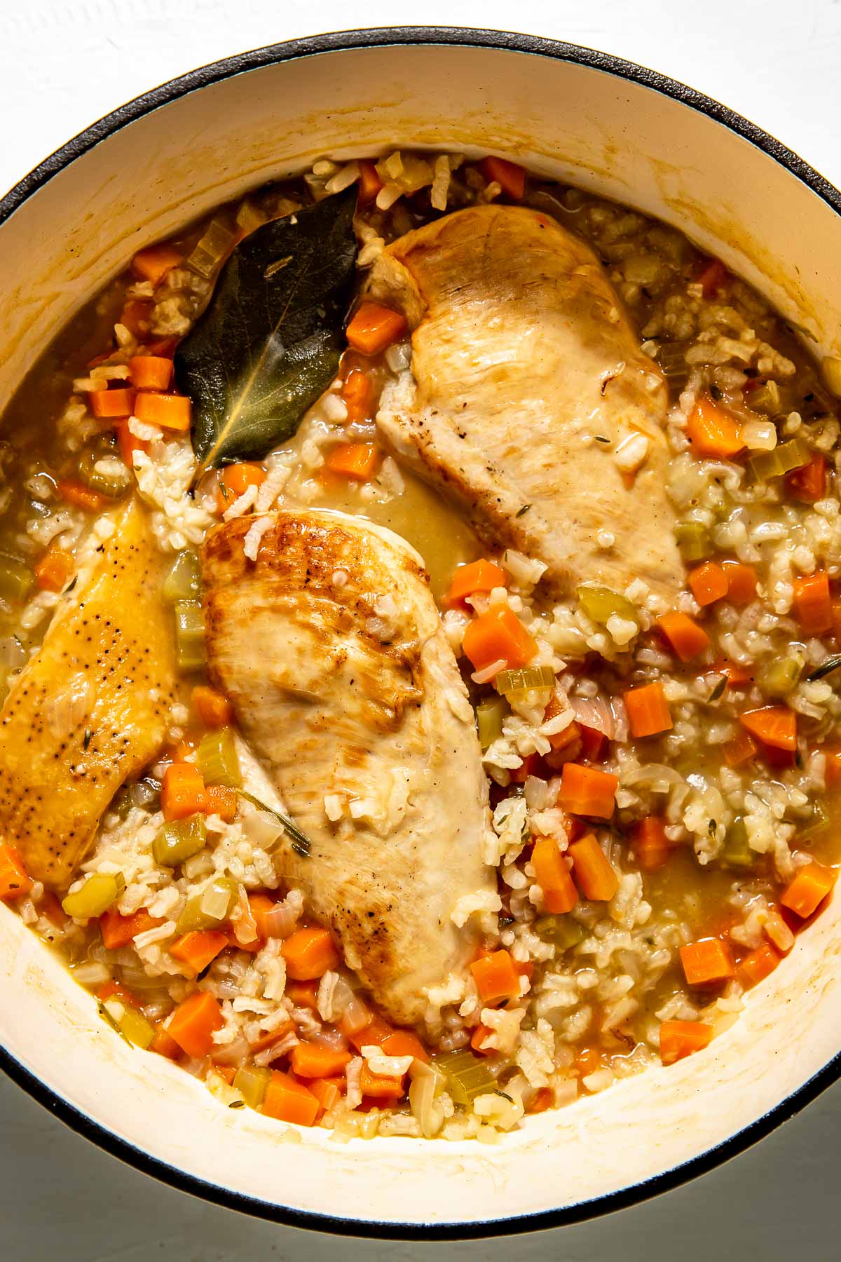 An overhead shot of simmering risotto with chicken breasts, a bay leaf and a parmesan rind in a large white pot atop a white textured surface.