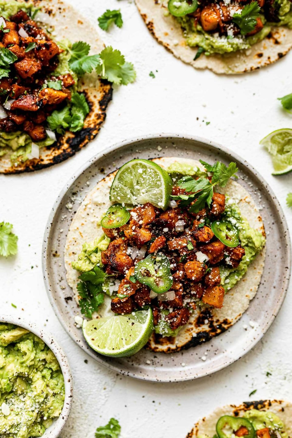 A chicken al pastor taco on a speckled ceramic plate, topped with thinly sliced jalapeno & cotija cheese. The taco is surrounded by 3 other al pastor tacos, a small bowl of mashed avocado, fresh cilantro leaves & lime wedges.