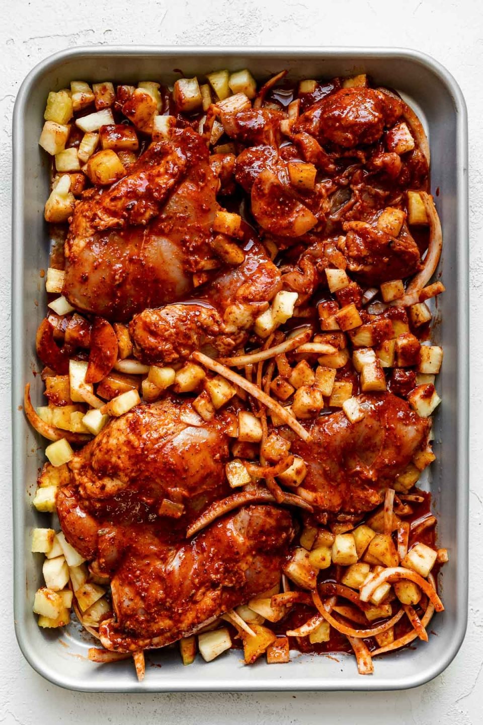 al pastor-marinated chicken thighs & sliced onion on a silver quarter baking sheet with finely diced pineapple. The sheet pan sits atop a white plaster surface.