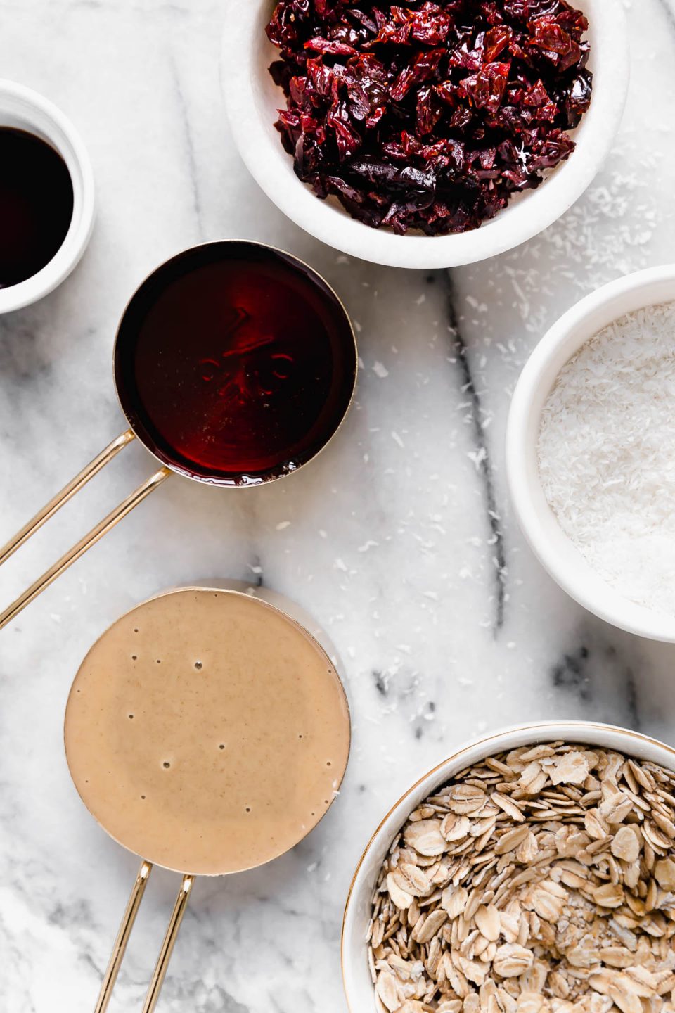 Vegan cookie dough bites recipe ingredients arranged on a white marble surface: tahini, rolled oats, shredded coconut, pure maple syrup, vanilla extract, & Montmorency tart cherries.
