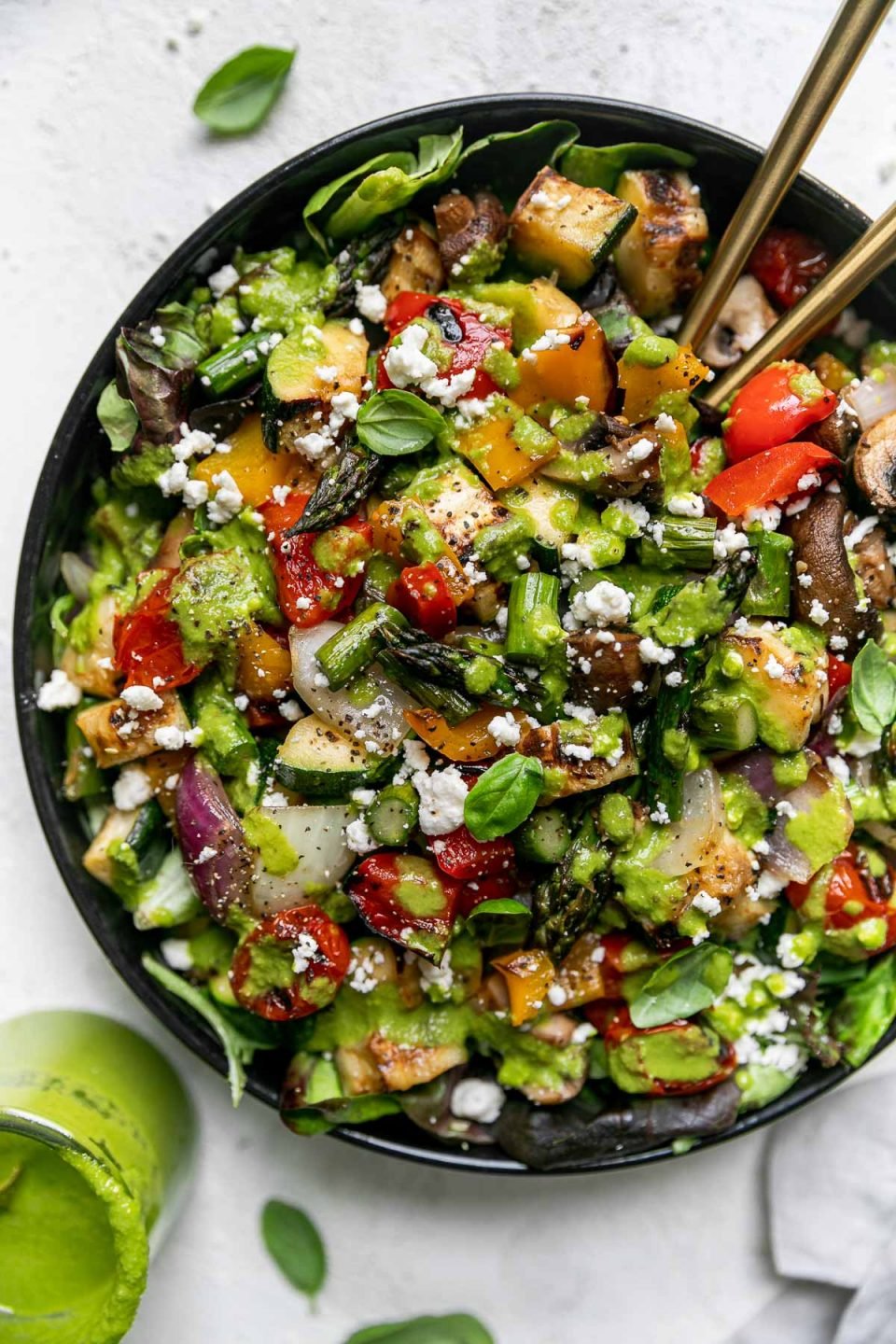 Chopped grilled vegetable salad in a large black serving bowl atop a gray linen napkin on a light gray surface. 2 gold serving spoons are nestled into the chopped vegetable salad. Fresh basil leaves and a jar of lemon basil vinaigrettes surround the serving bowl.