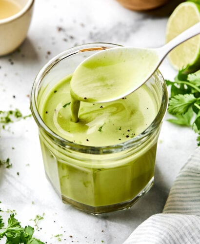 An angled shot of a clear glass of cilantro lime vinaigrette on a white marbled surface alongside cilantro leaves, salt and a halved lime. A spoon is drizzling dressing into the jar.