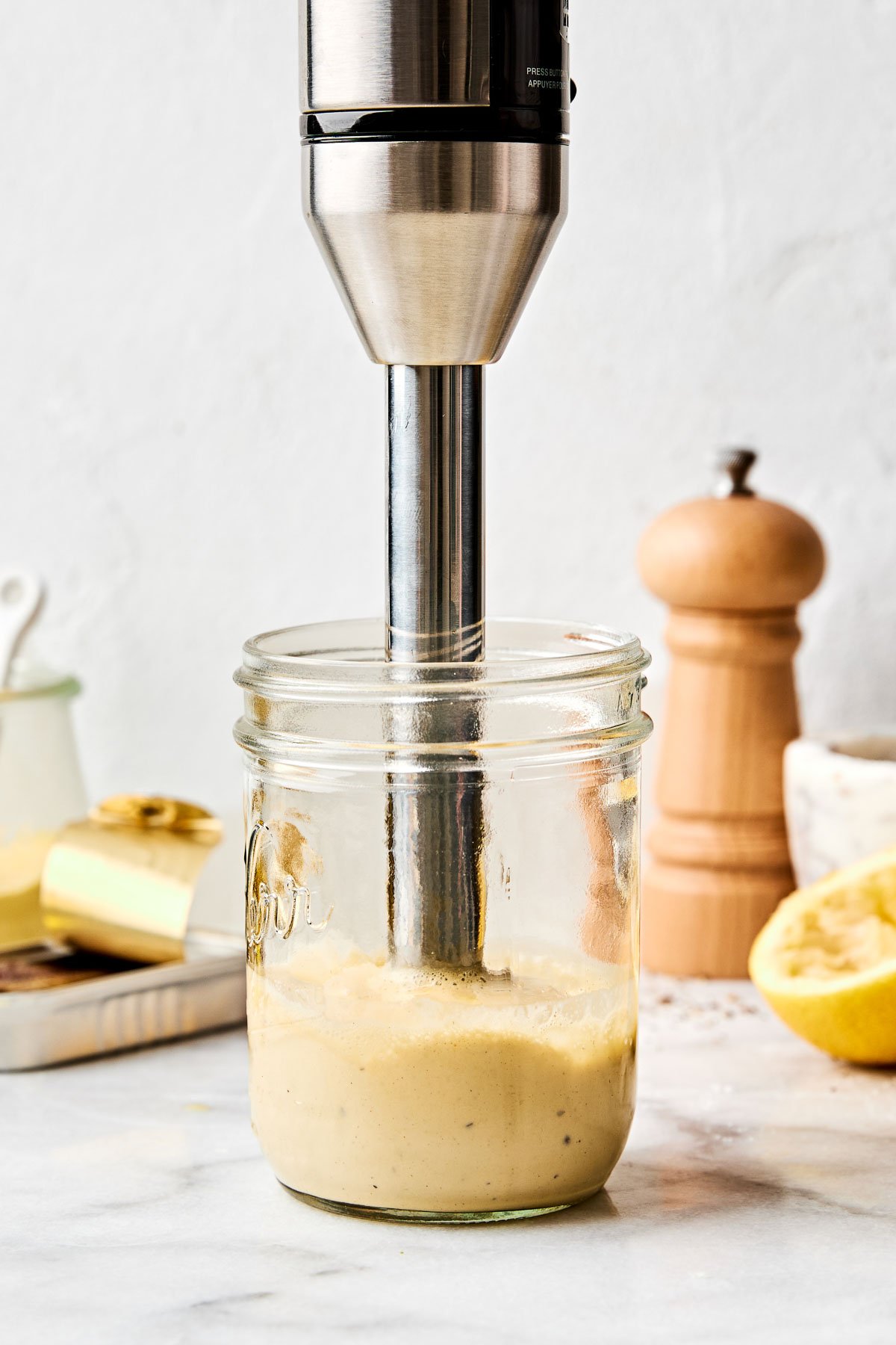 A side shot of an immersion blender blending Caesar dressing in a wide mouth jar atop a white marbled surface. A pepper mill, halved lemons, and dishes of ingredients sit in the background.