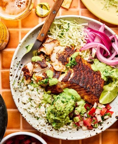 An overhead shot of a fish taco bowl with seasoned cod, rice, cabbage, red onion, guacamole, pico de gallo, and poblano ranch in a shallow stoneware bowl atop an orange tiled surface. Bowls of taco toppings sit alongside the bowl.