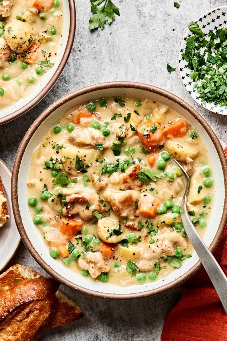An overhead shot of chicken stew topped with chopped fresh herbs in a stoneware bowl atop a grey speckled surface. The bowl is surrounded by a second bowl of stew, an orange cloth napkin, and slices of crusty bread.