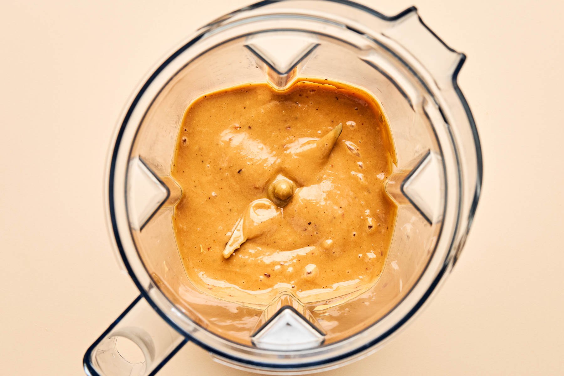 An overhead shot of peanut sauce in the bowl of a food processor atop a cream surface.