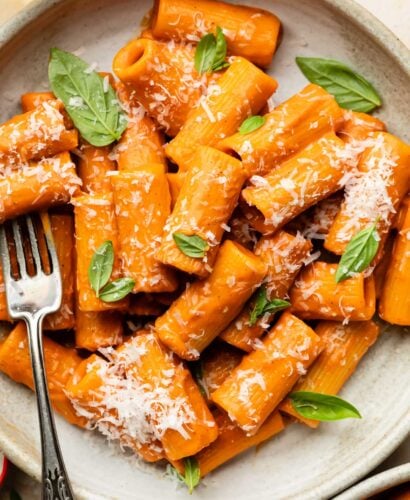 A plate of rigatoni pasta coated in creamy red pepper sauce, garnished with fresh basil leaves and grated Parmesan cheese, with a fork resting on the side.