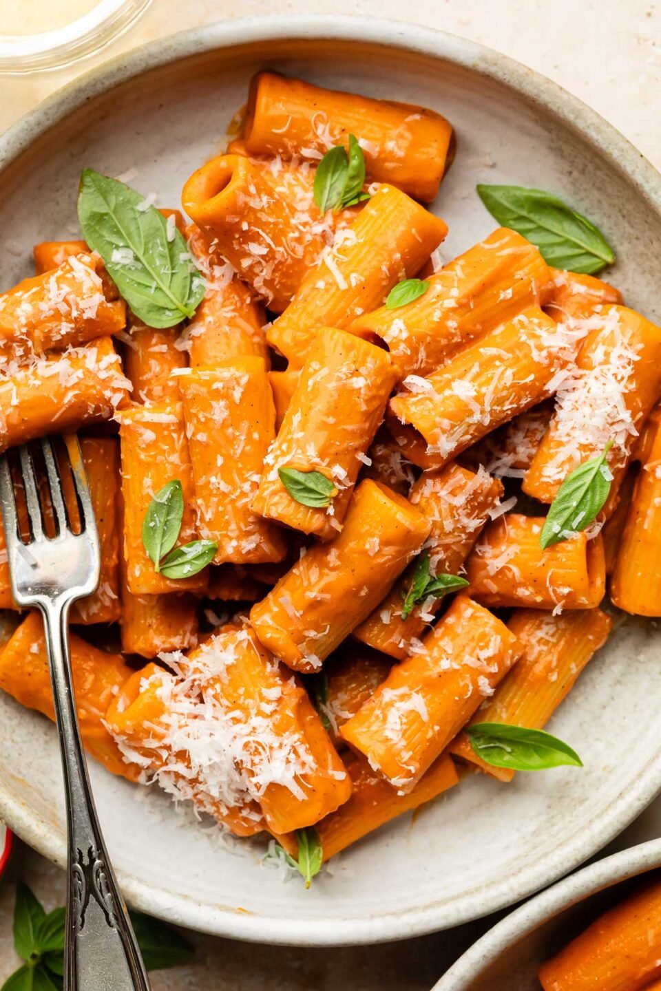 A plate of rigatoni pasta coated in creamy red pepper sauce, garnished with fresh basil leaves and grated Parmesan cheese, with a fork resting on the side.