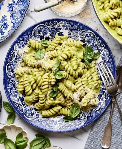 A blue and white plate filled with rotini pasta coated in creamy green sauce, garnished with fresh basil leaves and grated cheese. Surrounding the plate are utensils, a glass, and small bowls of grated cheese and basil.