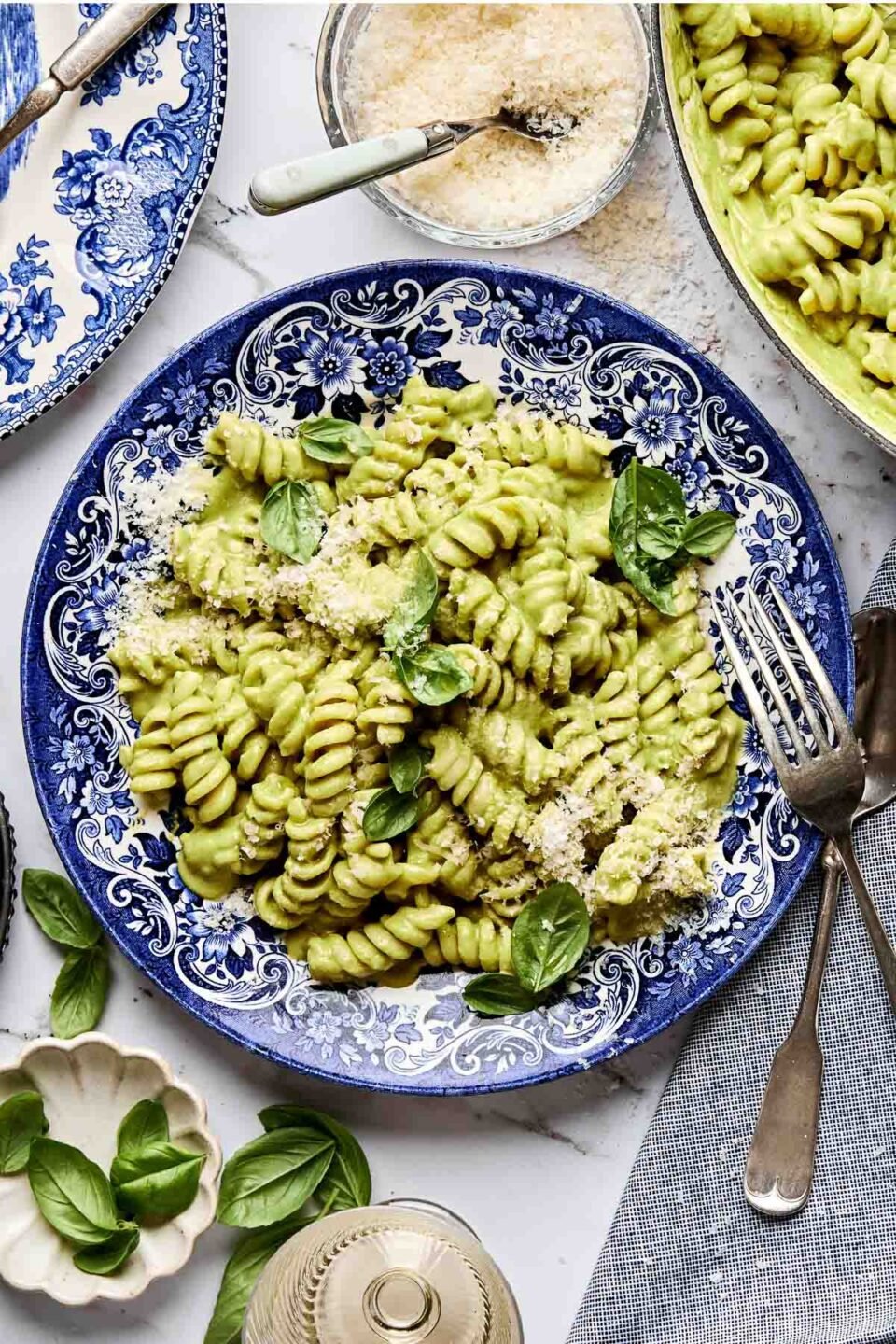 A blue and white plate filled with rotini pasta coated in creamy green sauce, garnished with fresh basil leaves and grated cheese. Surrounding the plate are utensils, a glass, and small bowls of grated cheese and basil.