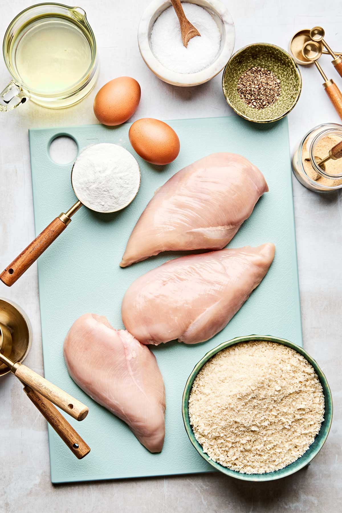 An overhead shot of ingredients on a light blue cutting board and a light grey textured surface: raw chicken breasts, panko breadcrumbs, salt, pepper, garlic powder, rice flour, eggs, and frying oil.
