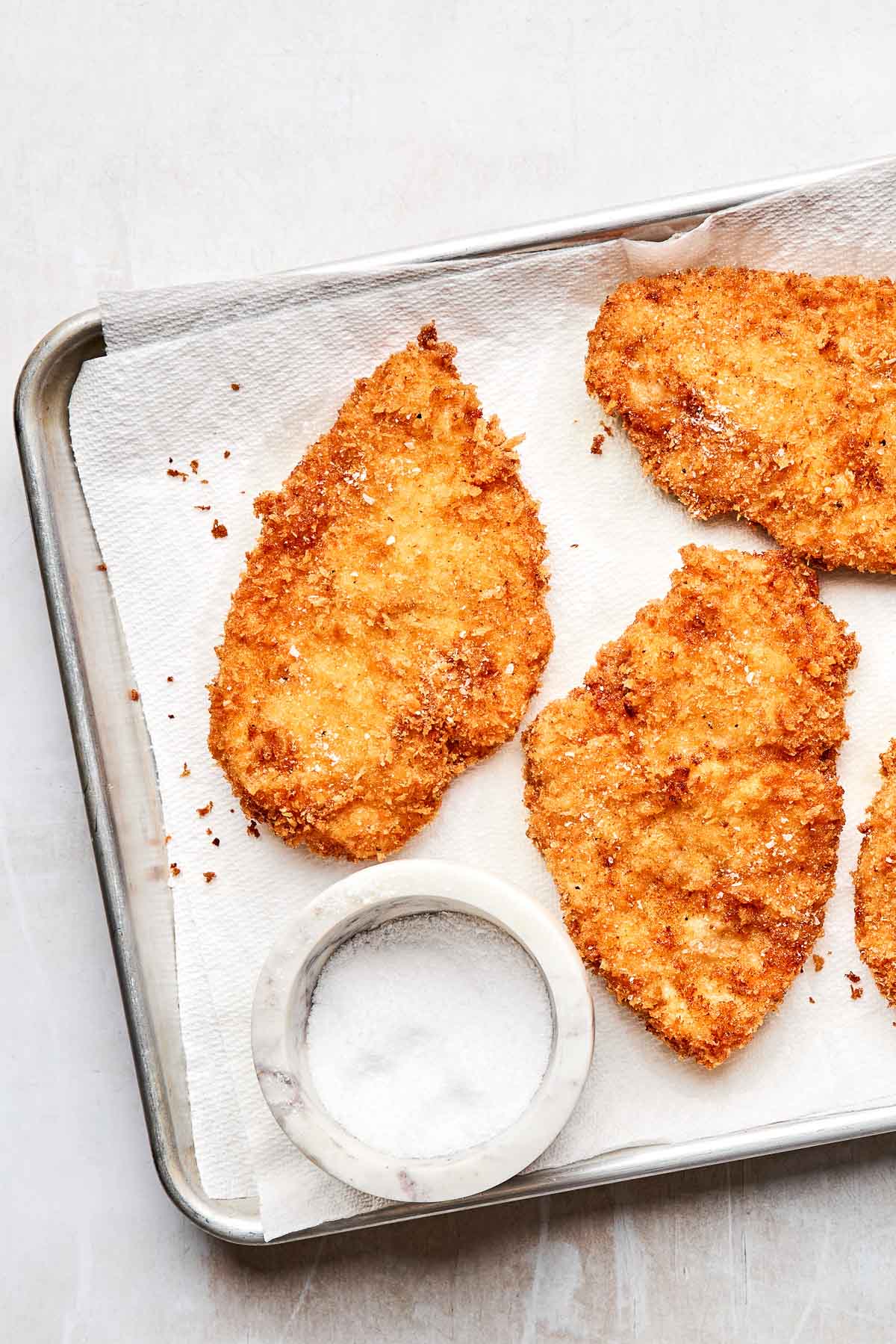 An overhead shot of four fried chicken breasts on a paper towel-lined sheet pan with a small dish of salt. The pan sits on a light grey textured surface.