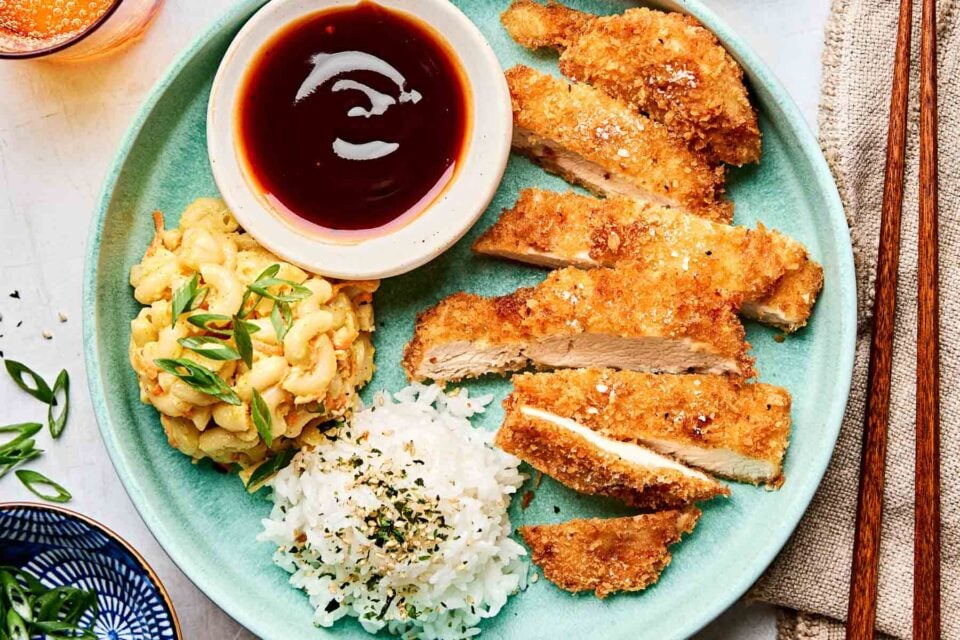 An overhead shot of sliced chicken katsu on a light teal plate alongside a small dish of katsu sauce, mac salad and white rice. The plate sits on a light grey textured surface with chopsticks and a bowl of sliced green onions.