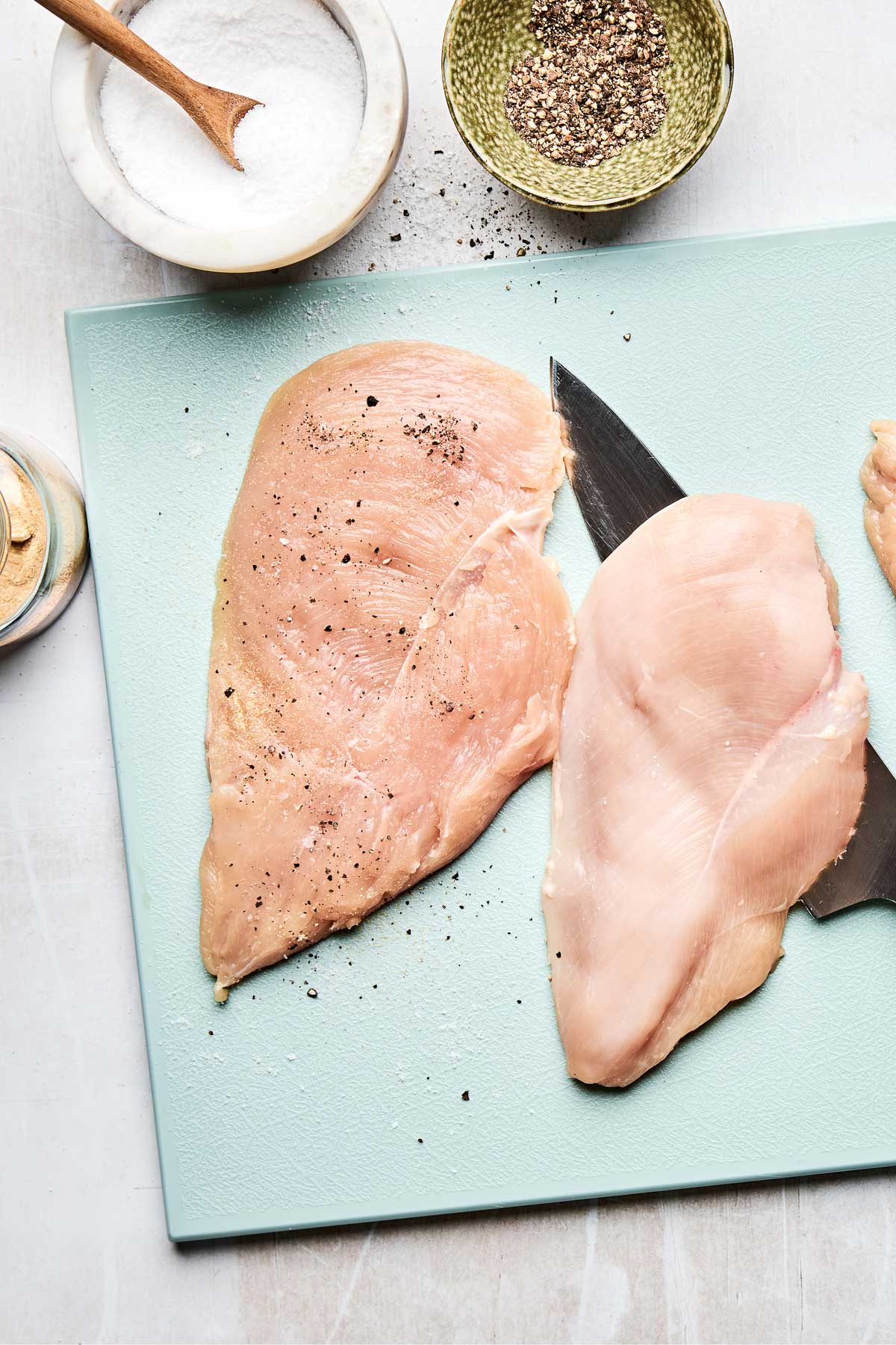 An overhead shot of butterflied chicken breasts with a chef's knife on a light blue cutting board atop a white textured surface. Dishes of salt, pepper and garlic powder sit alongside it.