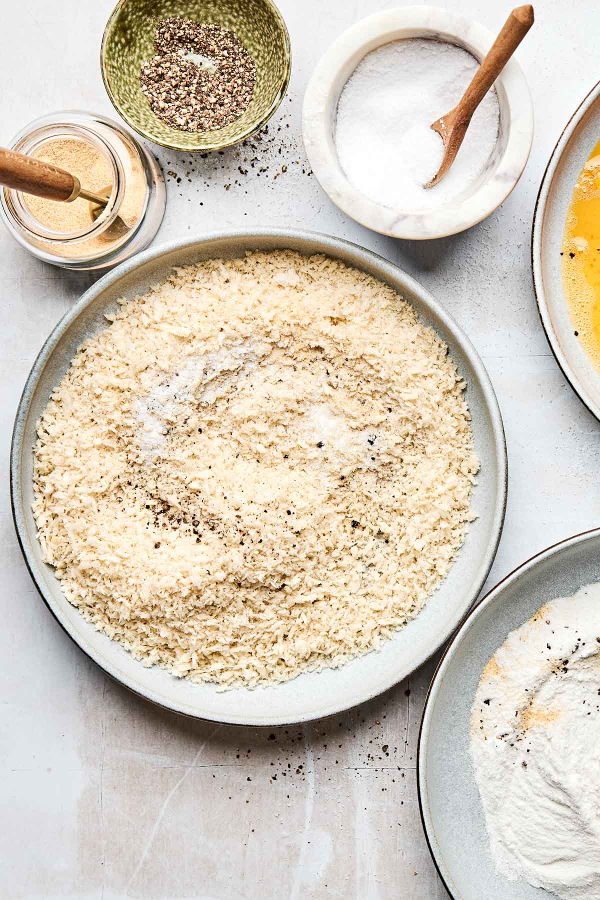An overhead shot of a dredging station: large bowls of breadcrumbs, flour, and eggs, alongside smaller dishes of salt, pepper, and garlic powder.