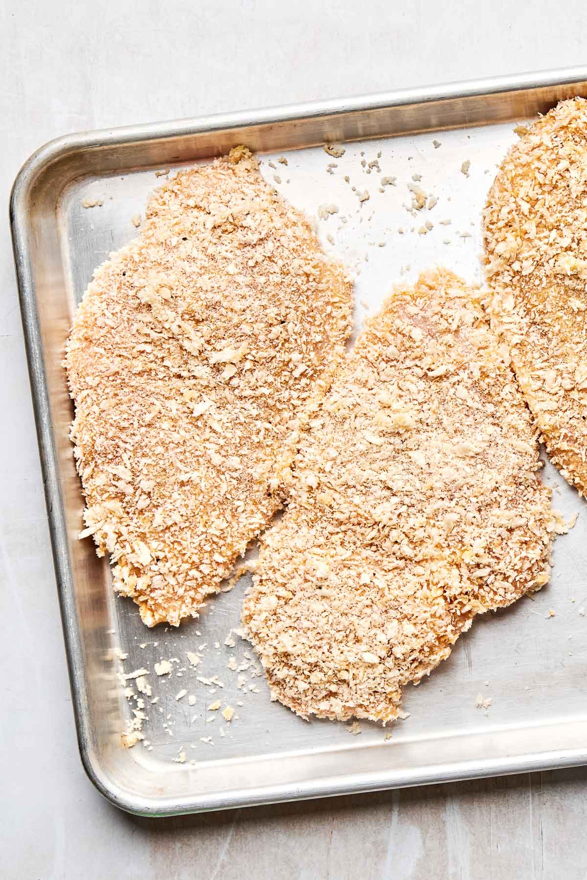 An overhead shot of breaded raw chicken breasts on a sheet pan atop a light grey textured surface.