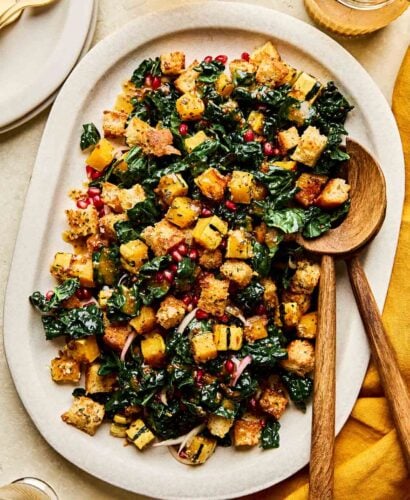An overhead shot of prepared fall panzanella salad on a white oval plate atop a textured beige surface. The tray sits alongside a golden cloth napkin, a jar of dressing, and glasses of white wine.